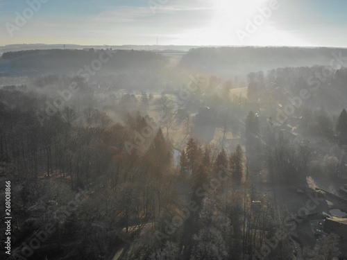 Aerial view of foggy and cold winter morning with blue sky facing the sun over the forest and farmland in Belgium  Walloon Brabant