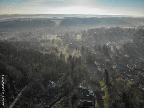 Aerial view of foggy and cold winter morning with blue sky facing the sun over the forest and farmland in Belgium, Walloon Brabant