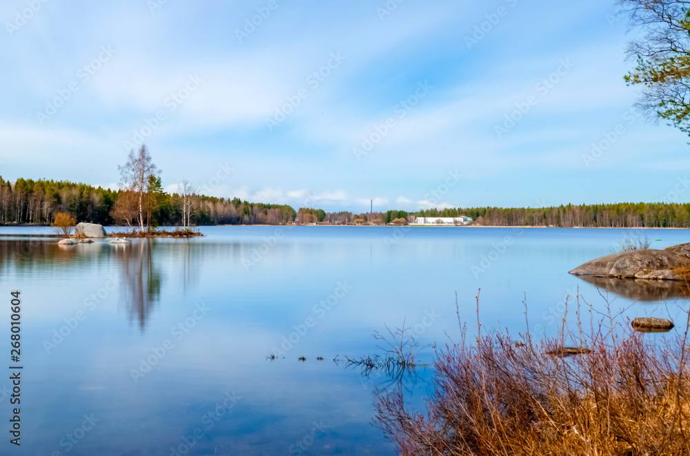 long exposure photograph of clean water lake surrounded by pine birch trees and scenic beauty under the cloud cover blue sky and reflection in water