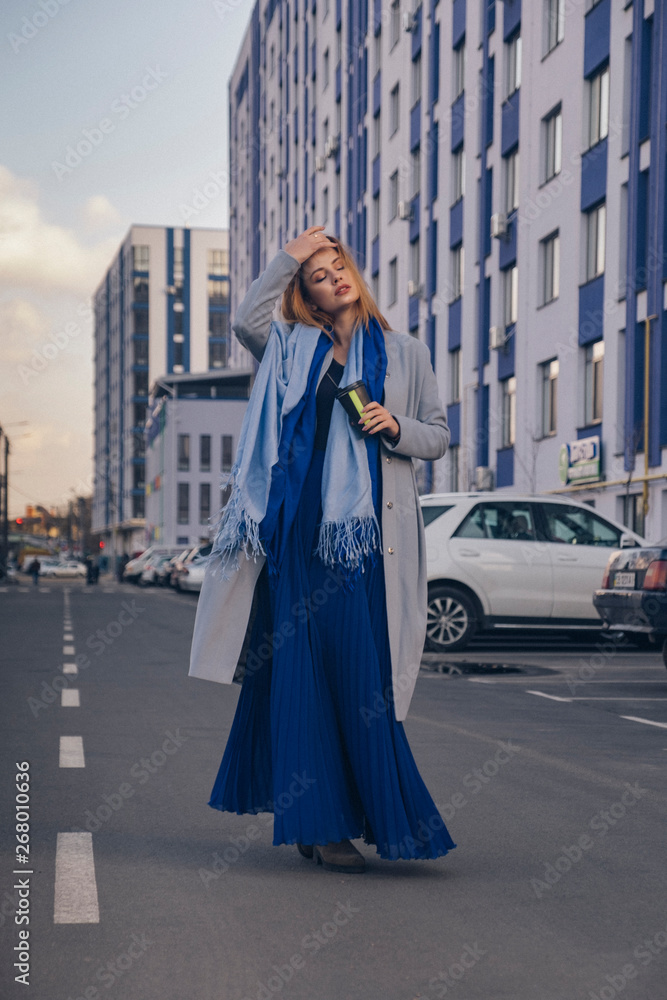 Fototapeta premium Gorgeous european woman in warm coat and dress on a walk in park near river. Windy weather. Her clothes fly in the wind. Sad, sensual and thughtful expression on her face