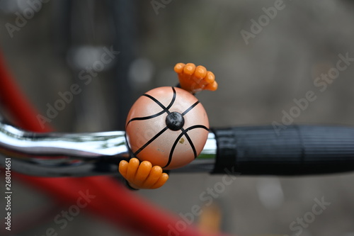 Two orange colored hands protecting a bicycle bell. An unusual bicycle bell in front of a blurred background.