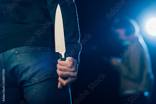 Foto cropped view of murderer hiding knife behind back on black