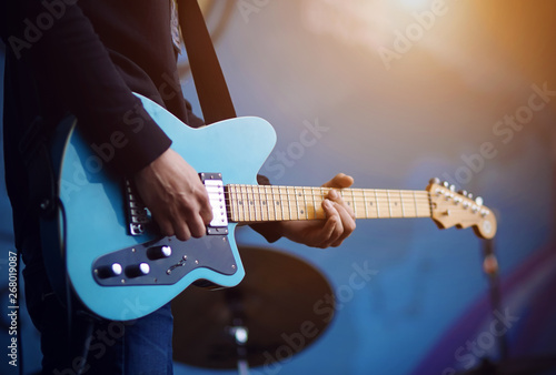 A man in a black sweater in blue jeans plays a blue electric guitar, illuminated by sunlight, on a blue background.