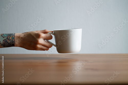 hand with tattoos holding a white mug on the table on a light background