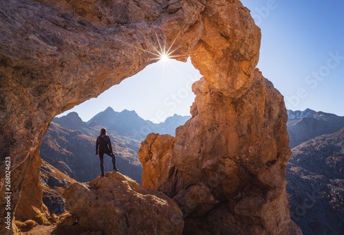 person standing on rock under arch formation rock while facing sun photo