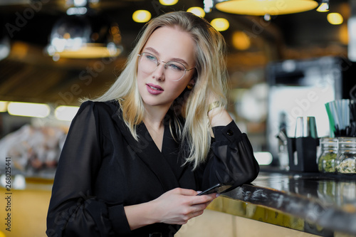 A young beautiful blond woman with glasses sits at the bar in a yellow interior. Drinks alcoholic cocktail with a straw and green mint.