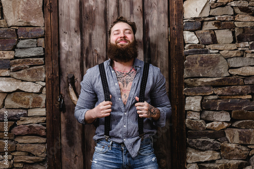 Funny brutal man with a beard and tattoos on his hands dressed in stylish casual clothes poses on the background of stone wall and wooden door © Leika production