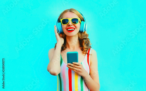 Portrait happy smiling woman holding phone listening to music in wireless headphones on colorful blue background