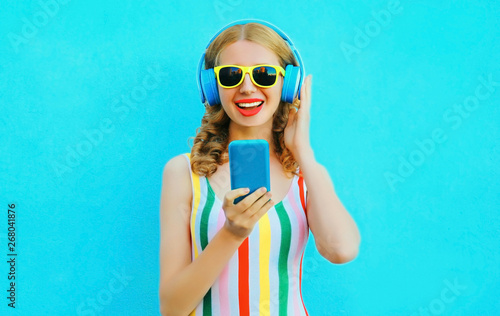 Portrait happy smiling woman holding phone listening to music in wireless headphones on colorful blue background