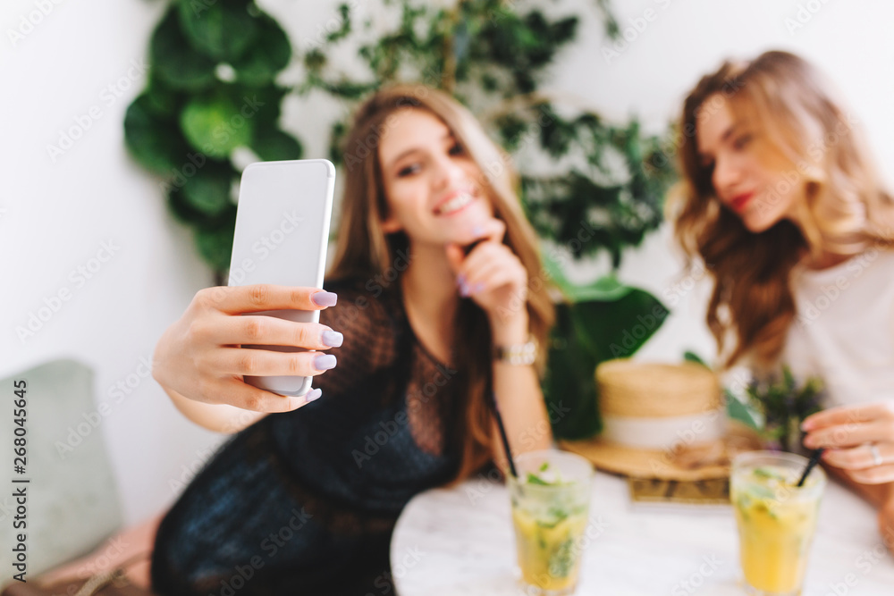 Gorgeous blonde young woman in stylish attire taking picture of herself on plant background while spending time with friend in cafe. Indoor portrait of two stylish girls enjoying juice in restaurant. Gorgeous blonde young woman in stylish attire taking picture of herself on plant background while spending time with friend in cafe. Indoor portrait of two stylish girls enjoying juice in restaurant.