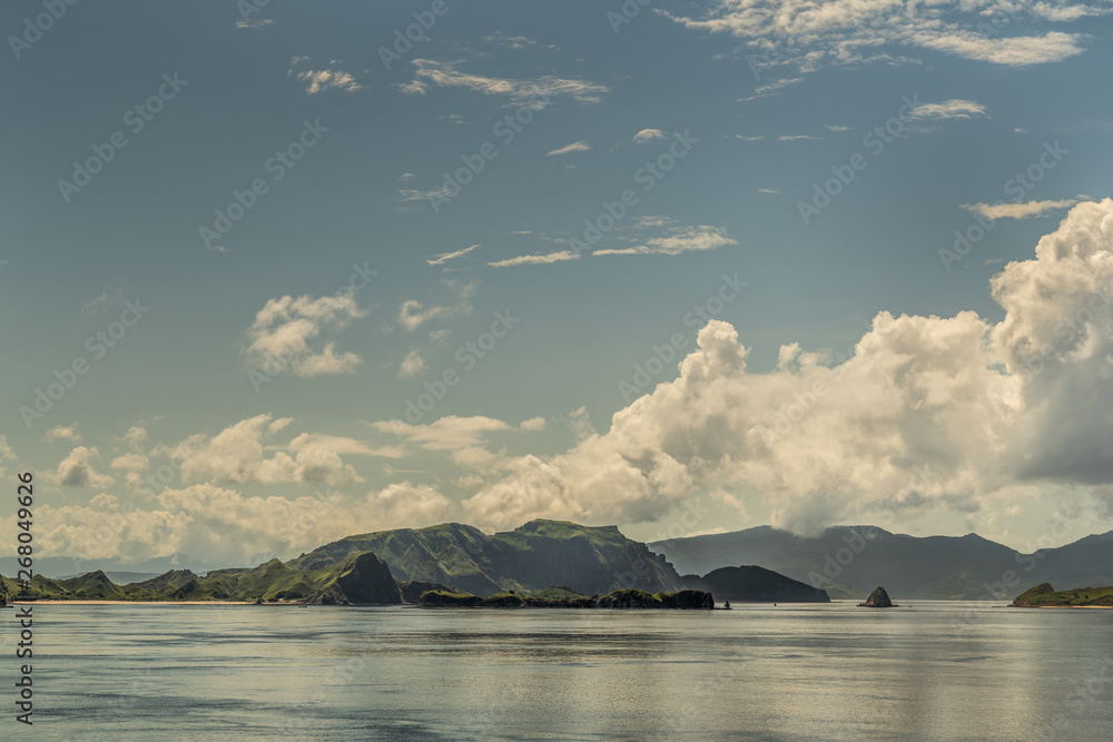 Rinca Island, Indonesia - February 24, 2019: Scenery of Westside coast in Savu Sea under cloudscape with white and darker patches. Green hills and smaller islands in front.