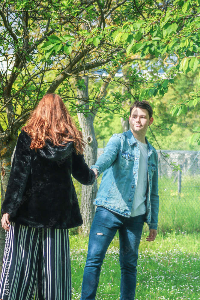 A handsome and young brown boy in jeans takes a girl with long red hair by the hand in nature on the grass