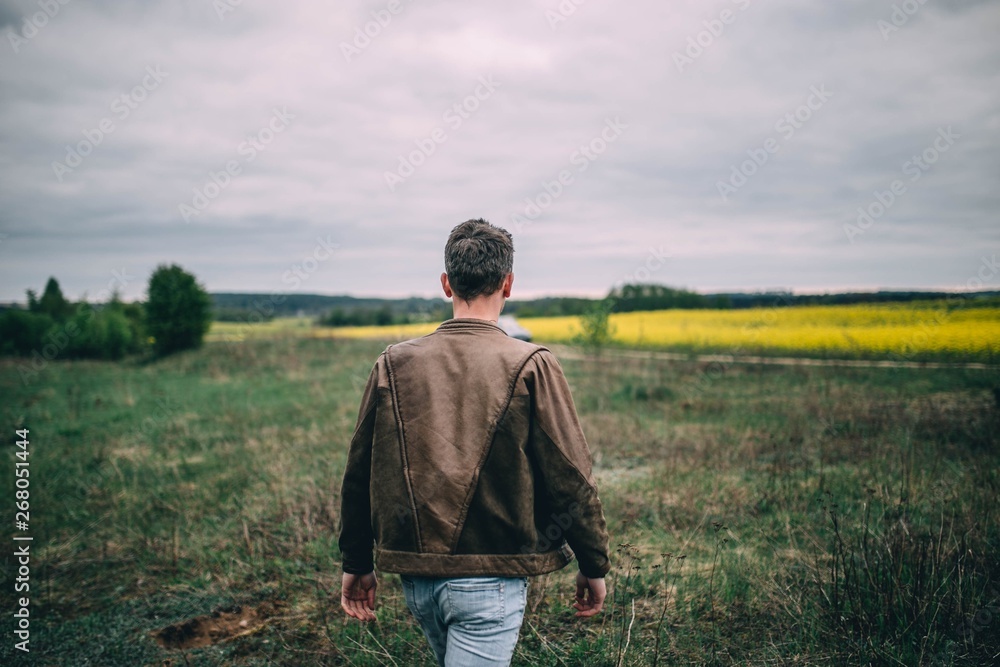 young lonely man in a leather brown jacket walks on the nature in the field