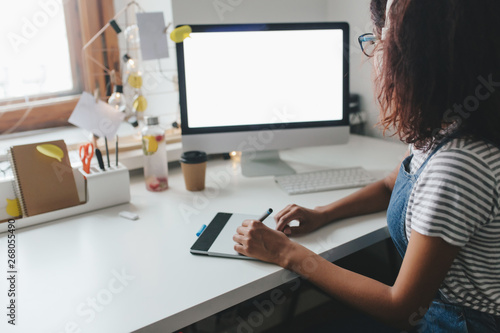 Brunette girl in striped shirt drawing on graphic tablet and looking at computer screen. Indoor photo of young woman with brown skin spending time in office, working on new project.