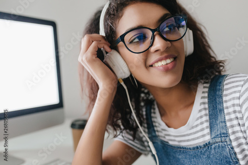Dreamy girl with shiny light-brown skin looking away gently smiling. Close-up portrait of curly brunette woman in glasses and headphones posing next to computer.