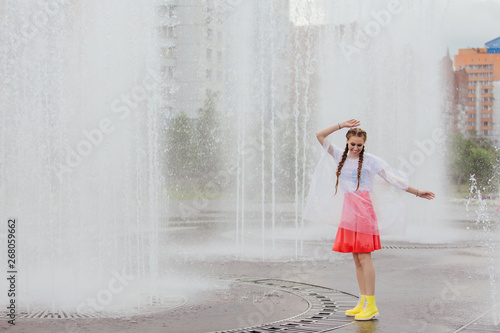 Young wet pretty girl with two braids in yellow boots stands near fountain.