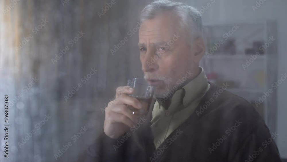 Retired male enjoying taste of whisky home, looking outside through rainy window Retired male enjoying taste of whisky home, looking outside through rainy window