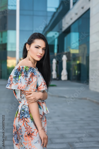 Young beautiful brunette woman with long hair in the street wearing white dress with flower pattern on the background of skyscraper © luckyphoto