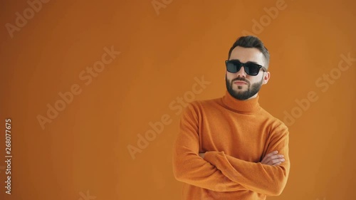 Portrait of confident bearded guy in sunglasses standing with asrms crossed and looking at camera against orange background. Cool people and style concept. photo