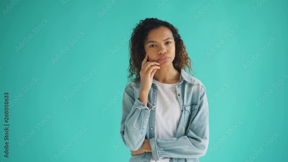 Portrait of thoughtful African American girl looking at camera on blue background thinking over problem making decision touching face. People and thoughts concept. Portrait of thoughtful African American girl looking at camera on blue background thinking over problem making decision touching face. People and thoughts concept.