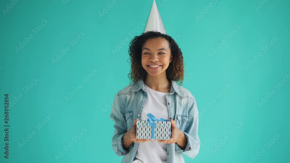 Portrait of attractive African American birthday girl in bright party hat holding gift box and smiling looking at camera. Happy young people and holidays concept. Portrait of attractive African American birthday girl in bright party hat holding gift box and smiling looking at camera. Happy young people and holidays concept.