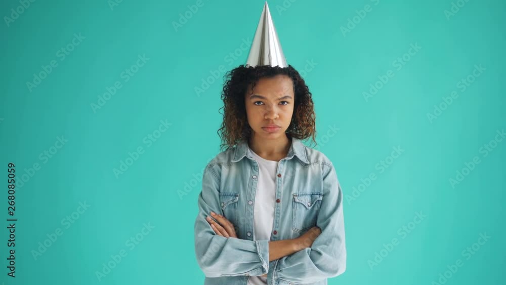 Portrait of unhappy mixed race girl in party hat standing with arms crossed and looking at camera with sad face. Bad mood, birthday and negativity concept. Portrait of unhappy mixed race girl in party hat standing with arms crossed and looking at camera with sad face. Bad mood, birthday and negativity concept.