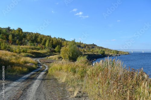 Road along the river to the forest on hills. Landscape in early autumn.