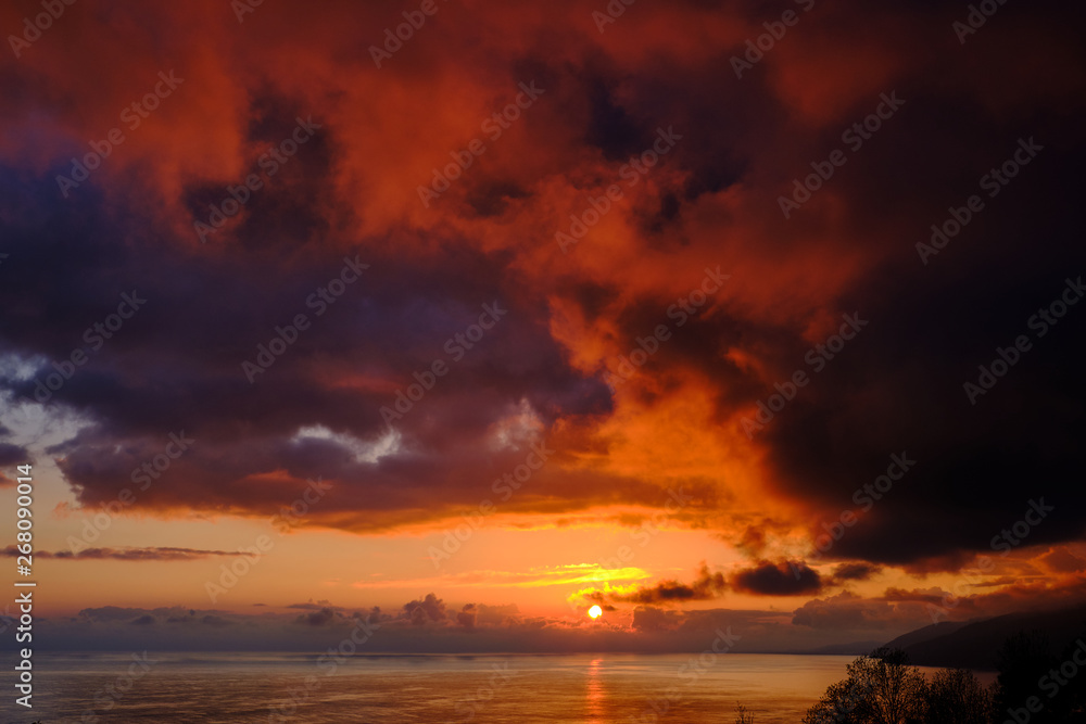 Sunset over the sea. Big orange clouds over mountains and water. Coastline. Storm off shore. Evening light.