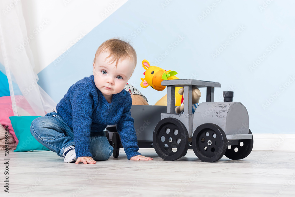 Boy blond in a blue sweater sits on a wooden floor. One year old baby playing with wooden toys. train made of wood, with girafe inside, blue wall at background Boy blond in a blue sweater sits on a wooden floor. One year old baby playing with wooden toys. train made of wood, with girafe inside, blue wall at background