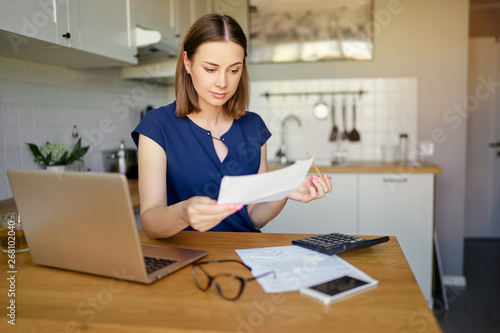 Thoughtful young woman using a laptop computer sitting at her kitchen holding utility bill and bank statements. Home interior.