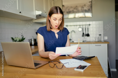 Stressed over bills. Portrait of surprised young woman using a laptop computer sitting at her kitchen holding utility bill and bank statements. Home interior. photo