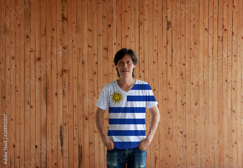 Man wearing Uruguay flag color shirt and standing with two hands in pant pockets on the wooden wall background.