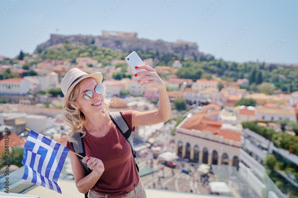 Enjoying vacation in Greece. Young traveling woman with national greek flag taking selfie on view of Athens city and Acropolis.