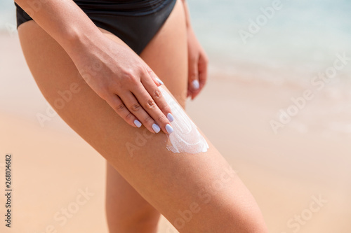 Woman in a black swimsuit is applying sun cream with her fingers on her leg at the sea background