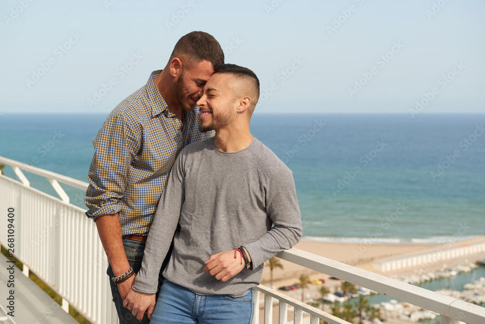 Una pareja gay en el balcón de su casa en un día soleado de verano con el puerto detrás.