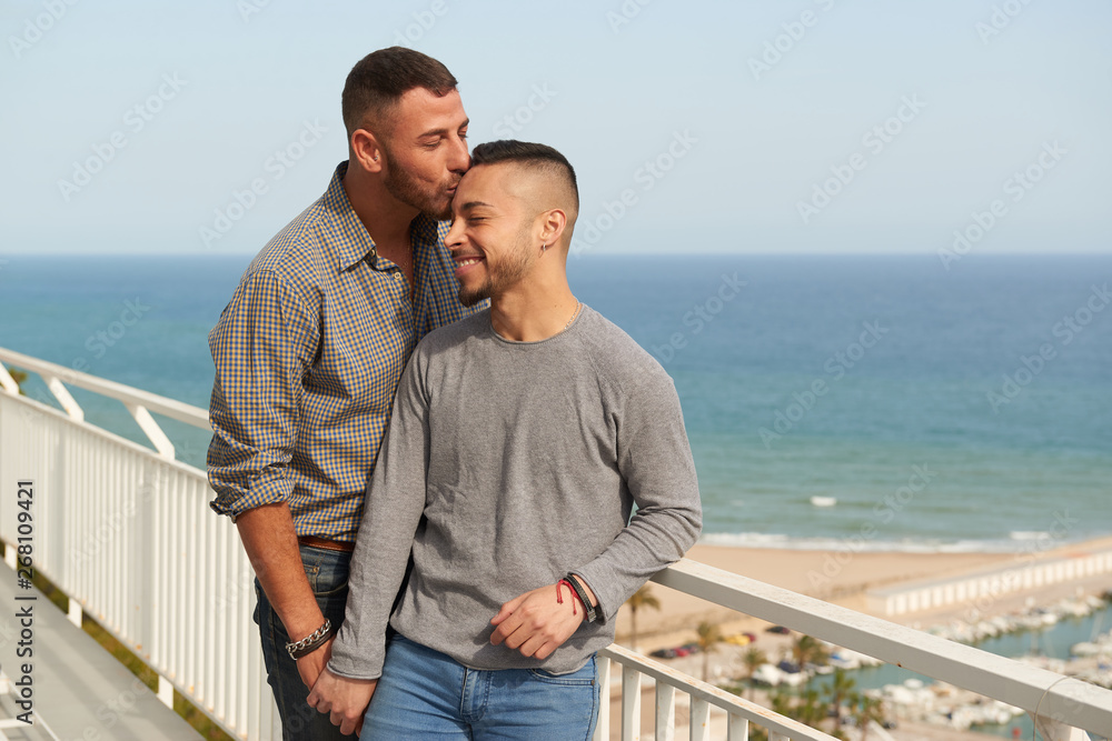 Una pareja gay en el balcón de su casa en un día soleado de verano con el puerto detrás.