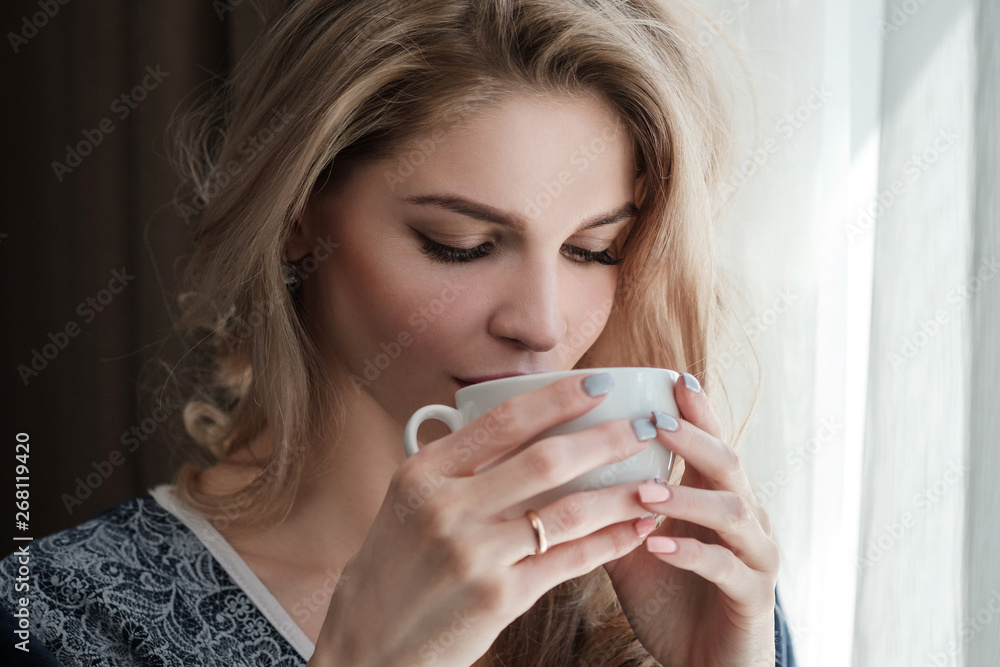 Beautiful young blonde woman in a blue robe by the window. Drinks coffee or tea from a white cup with a saucer. Morning, sunshine, bedroom window. Beautiful young blonde woman in a blue robe by the window. Drinks coffee or tea from a white cup with a saucer. Morning, sunshine, bedroom window.
