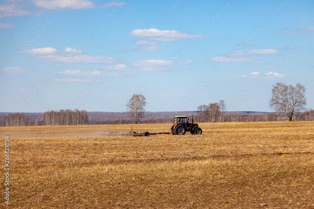 Fototapeta premium Red tractor with a trailed plow for mowing and weeding fields for the agro-industry of yellow color under the blue sky, a clear spring day. Preparation for planting crops rural technology. Farming.