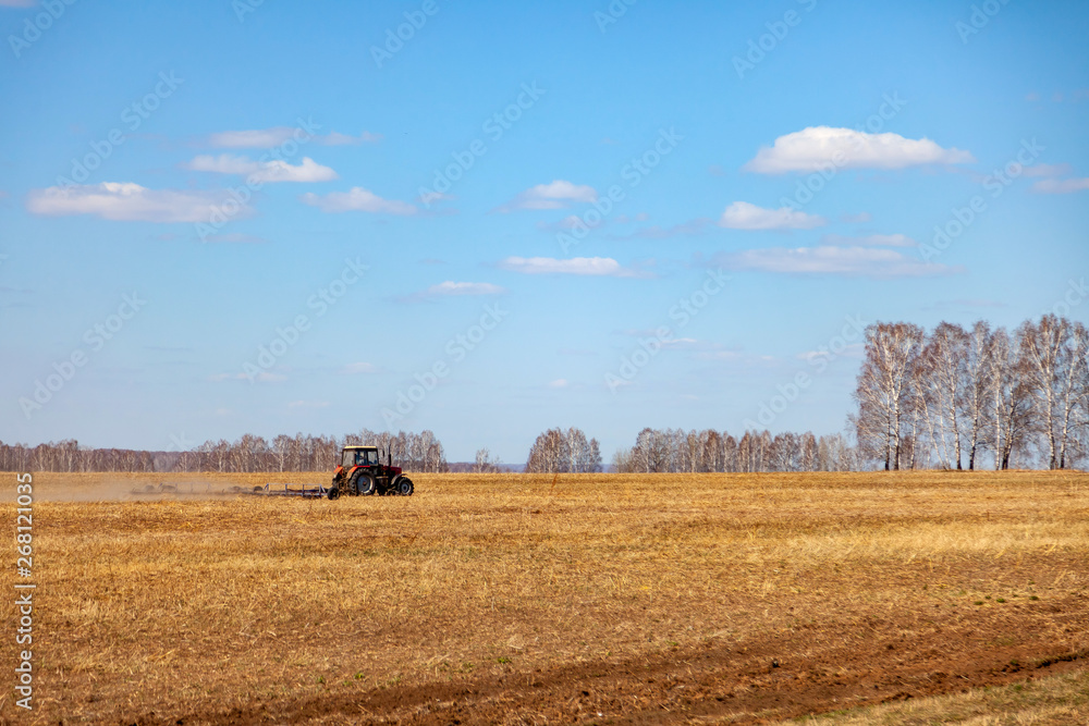 Red tractor with a trailed plow for mowing and weeding fields for the agro-industry of yellow color under the blue sky, a clear spring day. Preparation for planting crops rural technology. Farming.