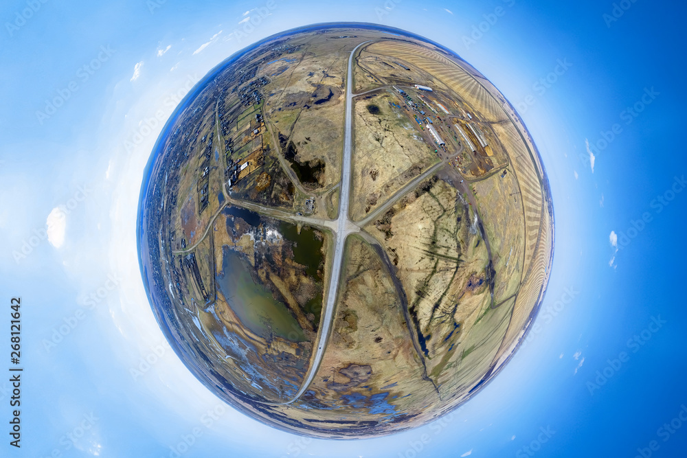 Aerial view of agricultural fields of yellow color crop under a clear blue sky with cloud on sunny warm spring day. Panorama 360 degrees. Little planet crossed by road with lake, crossroad and village