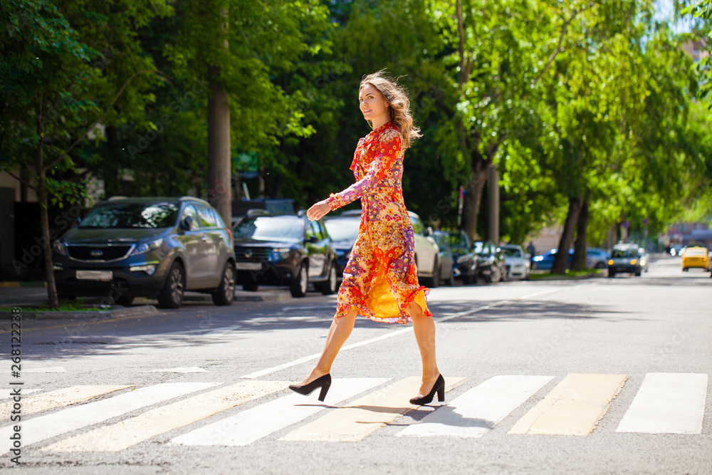 Young beautiful blonde woman in a red flower dress crosses the road at a crosswalk