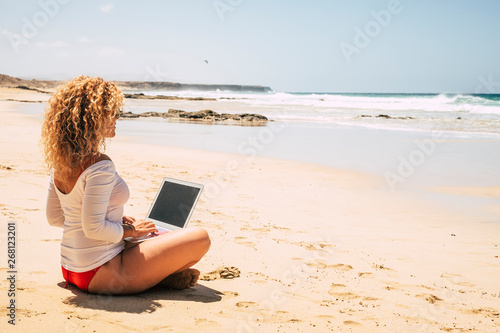 Concept of people work free from office - modern digital nomad lifestyle working in technology related jobs - happy women in bikini using laptop at the ebach enjoying the ocean view