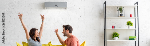 panoramic shot of excited man and woman sitting under air conditioner at home