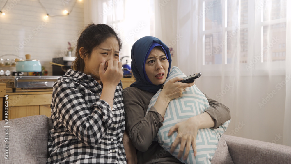 Scared multiracial young people watching thriller film on tv. two asian girl chinese and muslim sitting on couch with remote control hugging pillow surprised face with horror movie at home apartment. Scared multiracial young people watching thriller film on tv. two asian girl chinese and muslim sitting on couch with remote control hugging pillow surprised face with horror movie at home apartment.