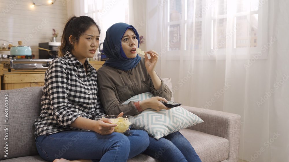 beautiful young asian women feeling thrilled while watching horror movie on television screen at home with bowl of potato chips. two arabic and chinese friends sitting on couch surprised emotion.