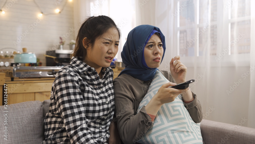 diverse female best friends sitting on couch with frowning face holding tv controller watching horror movie on television. two asian arabic and japanese roommates scared from thriller film hug pillow diverse female best friends sitting on couch with frowning face holding tv controller watching horror movie on television. two asian arabic and japanese roommates scared from thriller film hug pillow
