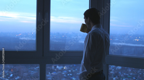 Young businessman drinking tea and admire view from window in the evening in office