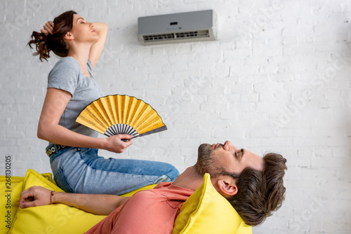 young woman with hand fan sitting on yellow sofa under air conditioner near lying man photo