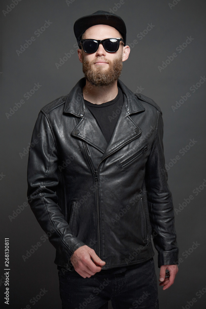 Plakat Man wearing leather biker jacket or asymmetric zip jacket with black cap, jeans and sunglasses. Handsome hipster man over gray background