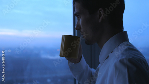 Young businessman drinking tea and admire view from window in the evening in office